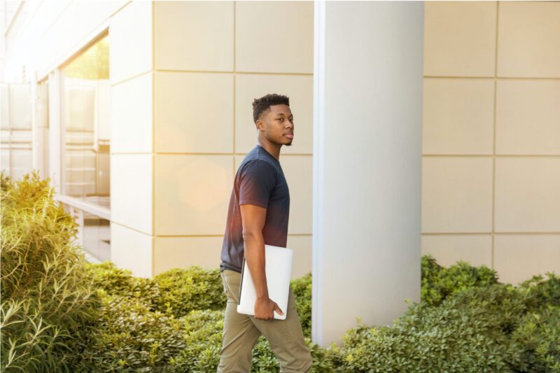 A male student walks toward a building holding a laptop